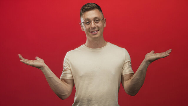 Young man wearing beige t shirt and glasses holding palms up and smiling against a red studio backdrop; playful indecision.