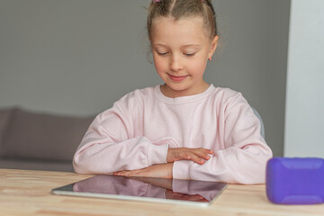 Naklejka premium A smiling young girl sitting at a wooden table looks down at a digital tablet.