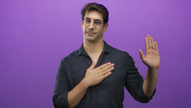Man wearing glasses and dark shirt, hand placed over chest and other hand raised in oath gesture in studio purple backdrop with wedding ring visible; sincerity.