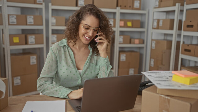 Woman talking on phone and working at laptop with parcels on desk in building; focus small business.