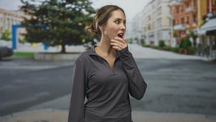 Fototapeta premium Woman covering mouth with hand on street while looking surprised, wearing dark gray zip pullover and bun hairstyle; surprise concern.