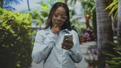 Fototapeta premium Woman holding smartphone with finger on chin and frowning while reading screen in studio; confusion troubleshooting.