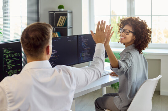Developers teammates highfive after coding success. At dual monitors in a bright office, coders celebrate solved tasks and smooth teamwork. Focused work. Strong concept of teamwork and success.