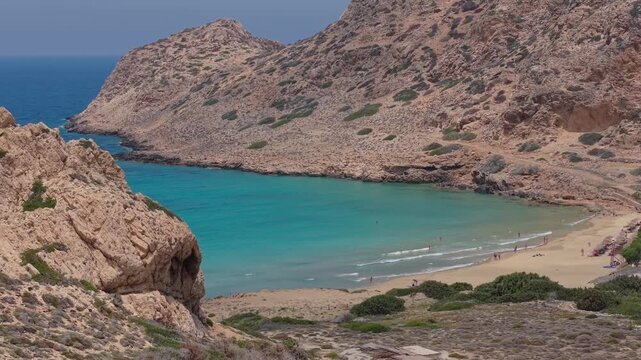 Drone footage revealing Agios Nikolaos Beach beyond the rocky terrain, with tourists enjoying the sandy shoreline and crystal-clear waters of Karpathos.