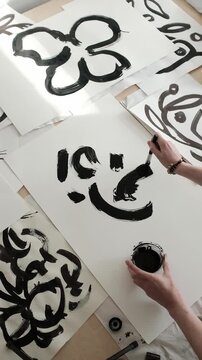 The artist holds a small container of black ink and uses a thick paint brush to draw fluid, sweeping strokes, forming a large abstract flower shape on a blank white sheet of paper. 