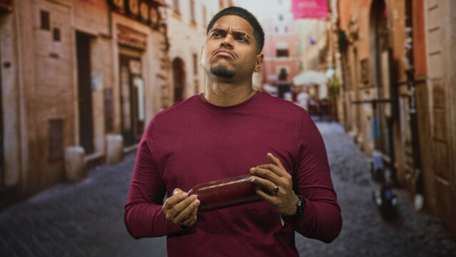 Man with beard holds a pomegranate juice bottle, shakes it with hands in a narrow street lined with old buildings; taste test disgust.