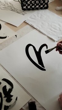 The artist holds a small container of black ink and uses a thick paint brush to draw fluid, sweeping strokes, forming a large abstract flower shape on a blank white sheet of paper. 