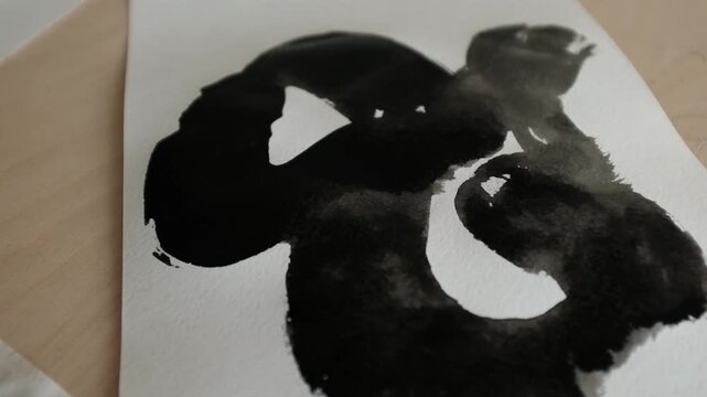 close-up shot capturing a hand using a round mop brush to create a bold, abstract ink painting on textured white paper, it absorbs the ink, showcasing realistic bleeding and dry brush strokes. 