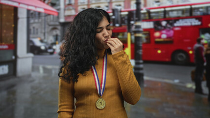 Fototapeta premium Woman wearing first place medal on rainy street in front of red doubledecker bus, hand raised near mouth with eyes closed; victory celebration pride.