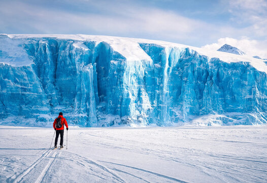 Lone cross-country skier in red jacket standing before a massive blue glacier ice wall, tiny person dwarfed by towering ice and snow