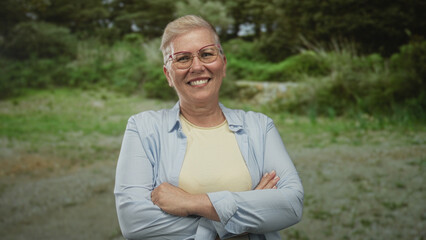 Obraz premium Middle aged woman wearing light blue shirt and yellow top, smiling with glasses and arms crossed in forest; calm confidence.