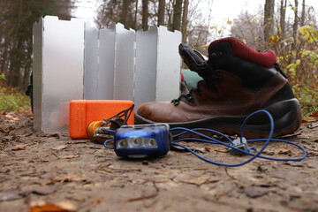 Horizontal view of camping gear on forest ground background including leather hiking boot, aluminum folding stove windshield screen, red headlamp flashlight, blue multi tool and orange foam pad © Krystsina