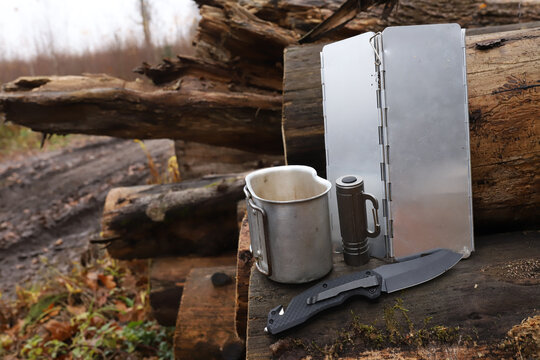 Horizontal view of bushcraft camping gear setup on old logs in forest including vintage aluminum military canteen cup with handles, tactical pocket folding knife and metal gas stove windshield screen