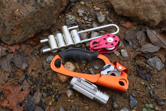 Top view flat lay of professional climbing gear on ground background including orange ascender handled rope clamp, red pulley, silver brake rack descender and led aluminum flashlight