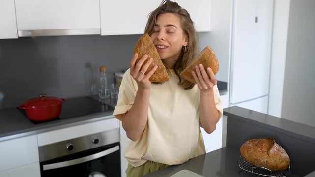 Young woman stands in modern kitchen, cutting freshly baked bread and enjoying its aroma. A housewife has baked sourdough bread in a cozy room. Concepts of cooking, comfort, and preparation.