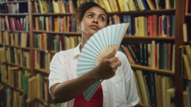 Woman fanning face with pale blue handheld fan in a library building while tilting neck and closing eyes; heat relief.