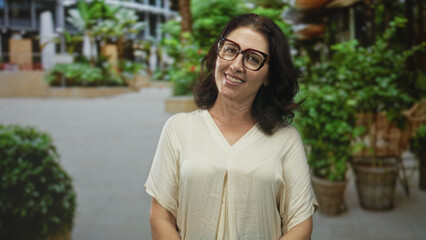 Fototapeta premium Middle aged woman smiles with face tilted up, wearing glasses in building atrium; quiet contentment.