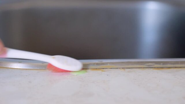 Closeup DIY grout hack using old toothbrush to scrape and lift grime at sink edge, fast strokes and targeted pressure remove stubborn film, relatable home remedy scene.