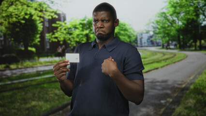 Fototapeta premium Man holding a credit card in left hand and points finger to card near his chest on a tree lined urban street with tram tracks; doubt identity security.