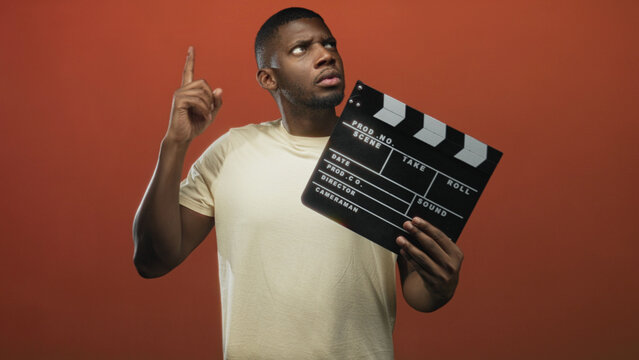 Man holding clapperboard and pointing index finger up in a studio with orange backdrop, wearing a light t shirt; curiosity idea.