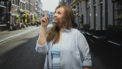 Fototapeta premium Senior woman pointing index finger and smiling on a narrow cafe street wearing light blue shirt and casual attire; friendly reminder.