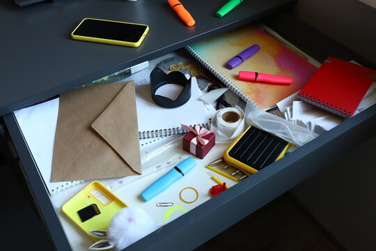 Top view of open messy office desk drawer filled with chaotic stationery items including kraft envelope, colorful highlighters, notebook, smartphone, sticky tape and small gift box for mess concept