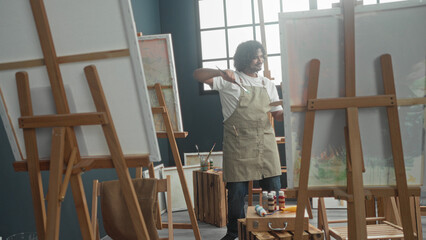 Fototapeta premium Young indian man painter in apron holding paintbrush and palette, raises brush to canvas in studio; serene concentration.