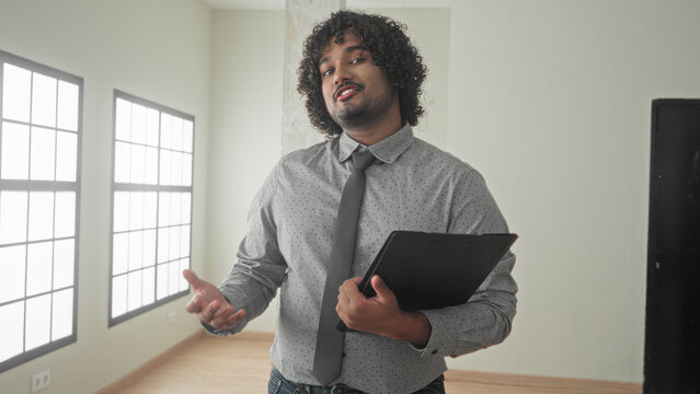 Young man holding clipboard and pointing finger in empty building interior with large windows and wooden floor; trust presentation confidence.