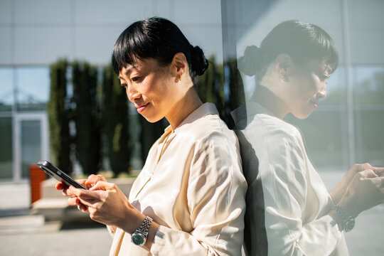 Businesswoman using smartphone by glass wall outdoors