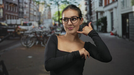 Fototapeta premium Woman with glasses making call me hand gesture on a city street lined with bicycles and brick buildings wearing a black long sleeve shirt; playful flirtation.