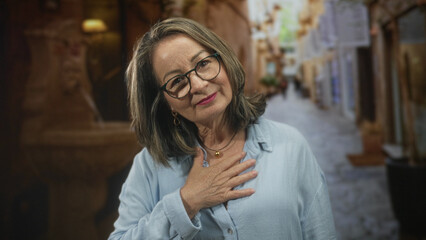 Fototapeta premium Senior woman with hand on chest, wearing glasses and necklace, standing in a narrow stone street; gratitude reflection memory.
