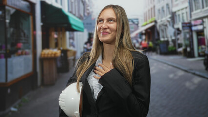 Fototapeta premium Young hispanic woman holding white hardhat waving hand on an urban street with storefronts; gratitude ambition.