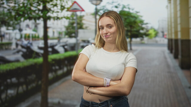 Woman with arms crossed showing bare midriff and wristwatch, smirking on street; confidence casual attitude.