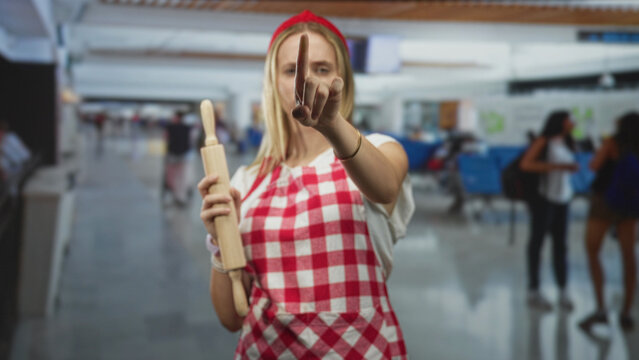 Woman chef in red checked apron holding rolling pin points index finger toward camera in airport terminal; assertive command.