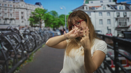 Fototapeta premium Woman wearing red glasses and white t shirt flashes two fingers by a canal on a street in amsterdam; playful confidence.