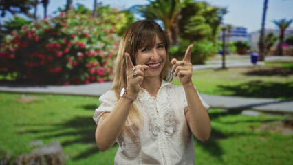 Fototapeta premium Woman pointing both index fingers up while smiling in a park by a path and flowering shrub near palm trees; playful.