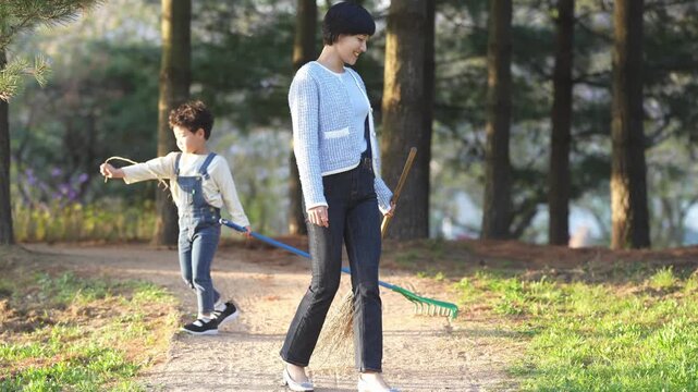 Soft sunlight bathes a park path lined with tall trees. Woman walks ahead, holding a broom. Her smile shows quiet contentment. Child runs beside her, laughing, with a stick