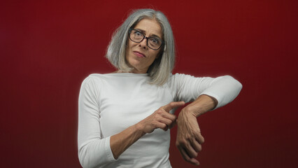 Fototapeta premium Middle age grey haired woman in white shirt pointing finger to wrist in studio with red wall; time impatience.