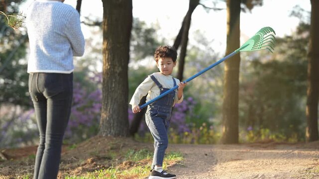 Child rakes dirt near trees with adult nearby. Sunlight glows through branches overhead. Child smiles, happy, focused on task. Natural setting, quiet, peaceful afternoon. Great for family outdoor