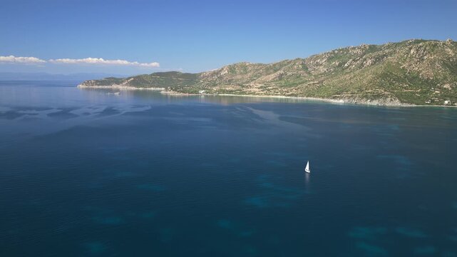 Aerial drone footage of a lone sailboat gliding across calm blue Mediterranean waters off the rugged coastline of Villasimius, Sardinia, with mountains and shoreline in the background