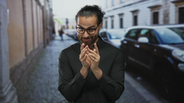 Man with glasses rubbing fingers together in black shirt on cobblestone street; scheming slyness cunning intent.