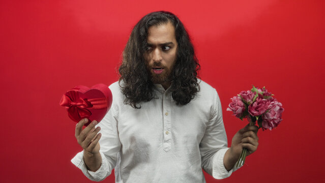 Man holding a red heart box and pink bouquet in a red studio while grimacing and offering gifts; confusion.