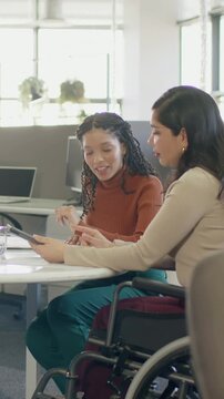 Vertical video: Offering tablet, beige blazer wheelchair holding rust top peer reviewing at office