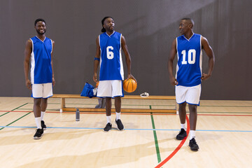 Naklejka premium African American male players wearing blue jerseys standing on court holding basketball near bench
