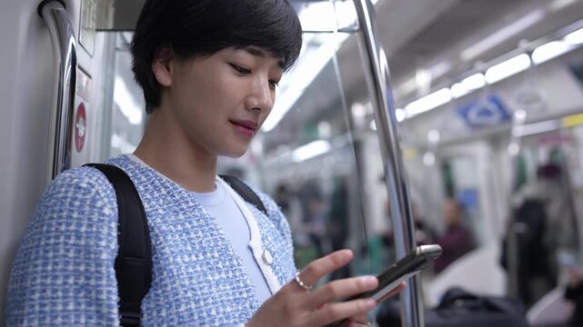 Young woman rides subway, focused on phone. She wears blue patterned jacket, carries black backpack. Subway interior shows soft lighting, metal poles, blurred passengers