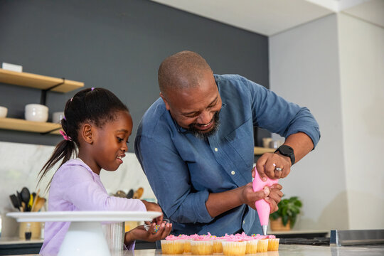 African American father and child decorating cupcakes at kitchen counter using pink piping bag