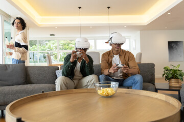 Obraz premium Diverse teenage males playing VR games on gray sofa wearing white headsets and holding controllers
