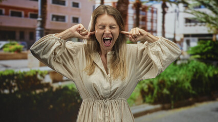Fototapeta premium Woman plugging ears with fingers and open mouth shouting on a street near apartment buildings and hedges; annoyance overload.