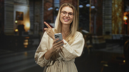 Fototapeta premium Blonde woman holding smartphone points finger and smiles in an ornate museum building, wearing glasses and beige linen dress; curiosity.