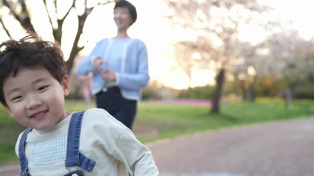 Soft sunlight glows behind cherry blossoms. Woman smiles warmly as child runs toward her. Child laughs joyfully, arms held wide. Perfect spring scene for family moments or happy memories